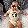 baby, sunglasses, onesie, couch, cushion, infant, child, text_on_clothing, indoor, relaxing, portrait, person, comfortable, fashion, cute, skin, plush, seated, expressionless, home