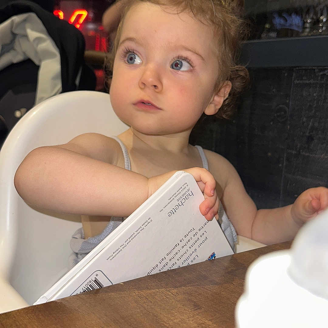 Luna participe au concours pour gagner de l'argent avec cette photo : baby, background, blue_eyes, blurry, book, child, clothing, curious, curly_hair, face, high_chair, holding, indoor, person, reading, restaurant, seated, table, toddler, wooden_table