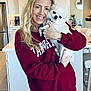 blonde_hair, cabinet, casual_clothing, chair, counter, decor, dog, harvard, holding, indoor, kitchen, light_wood_floor, pet, portrait, red_sweatshirt, refrigerator, smile, table, tiara, woman