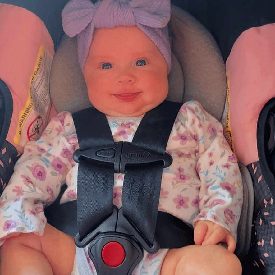 Sofia is registered to the contest to win money with this photo: baby, car_seat, child, smiling, headband, floral_clothing, safety_harness, pink, grey, infant, seatbelt, portrait, indoors, cute, happy, sitting, child_seat, car, young_child, closeup
