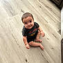barefoot, cabinet, casual, child, clothing, curious, face, floor, home, indoor, light, looking_up, person, shorts, sitting, smiling, toddler, tshirt, wooden_floor, young_child
