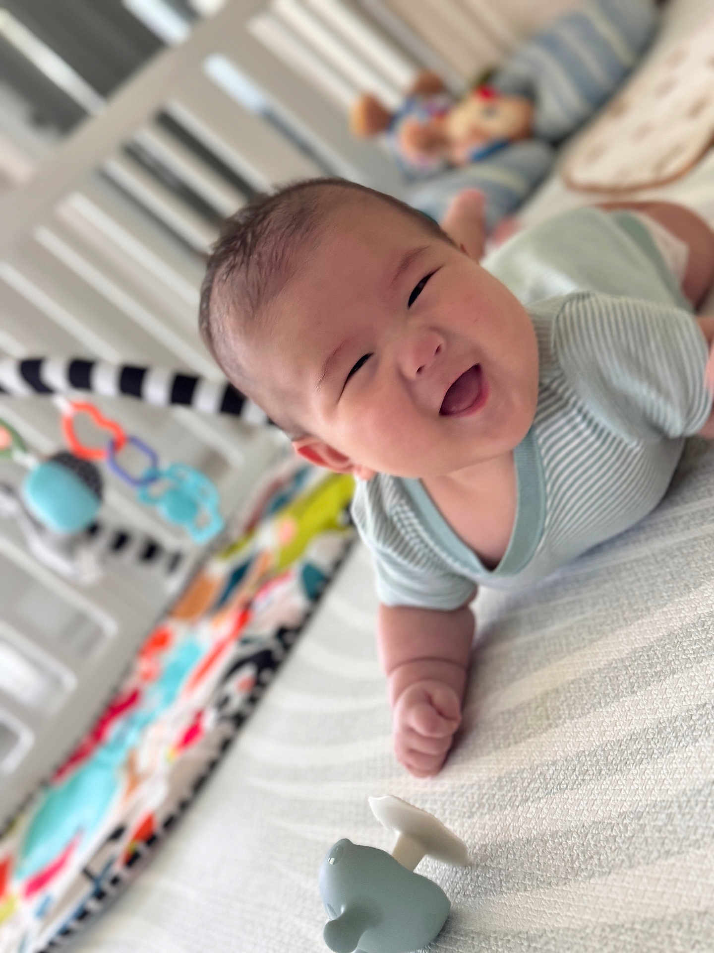 Ean is registered to the contest to win money with this photo: baby, smiling, tummy, toy, crib, colorful, happy, infant, clothing, playtime, child, indoors, cute, face, person, soft, blanket, striped, pattern, comfort