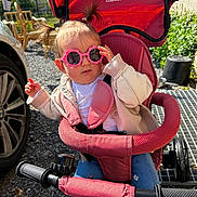 Eva participe au concours pour gagner de l'argent avec cette photo : toddler, child, sunglasses, pink, pigtails, stroller, outdoor, sunny, casual_clothing, jeans, jacket, toy_bike, wheel, gravel, car, hand, face, seat, nature, shade