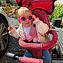 toddler, child, sunglasses, pink, pigtails, stroller, outdoor, sunny, casual_clothing, jeans, jacket, toy_bike, wheel, gravel, car, hand, face, seat, nature, shade
