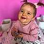 baby, child, girl, smile, pigtails, pink_bow, pink_background, holding_object, happy, portrait, cute, indoor, clothing, long_sleeves, patterned_blanket, face, eyelashes, skin, cheeks, toy