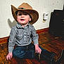 child, toddler, cowboy_hat, shirt, jeans, boots, floor, wooden_floor, indoor, sitting, smiling, curious, shadow, wall, plaid, patterned_clothing, young_child, cute, portrait, casual