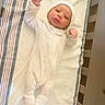 Jose is registered to the contest to win money with this photo: baby, newborn, infant, onesie, hat, blanket, crib, striped, hand, face, sleepwear, footwear, soft, cute, lying_down, person, child, indoors, resting, cozy