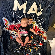 Evelyn joined the competition — help win amazing prizes! baby, infant, onesie, blanket, racing_theme, chair, indoor, person, clothing, expression, sitting, colorful, pattern, text_on_clothing, paper, notebook, furniture, curious, cute, young_child