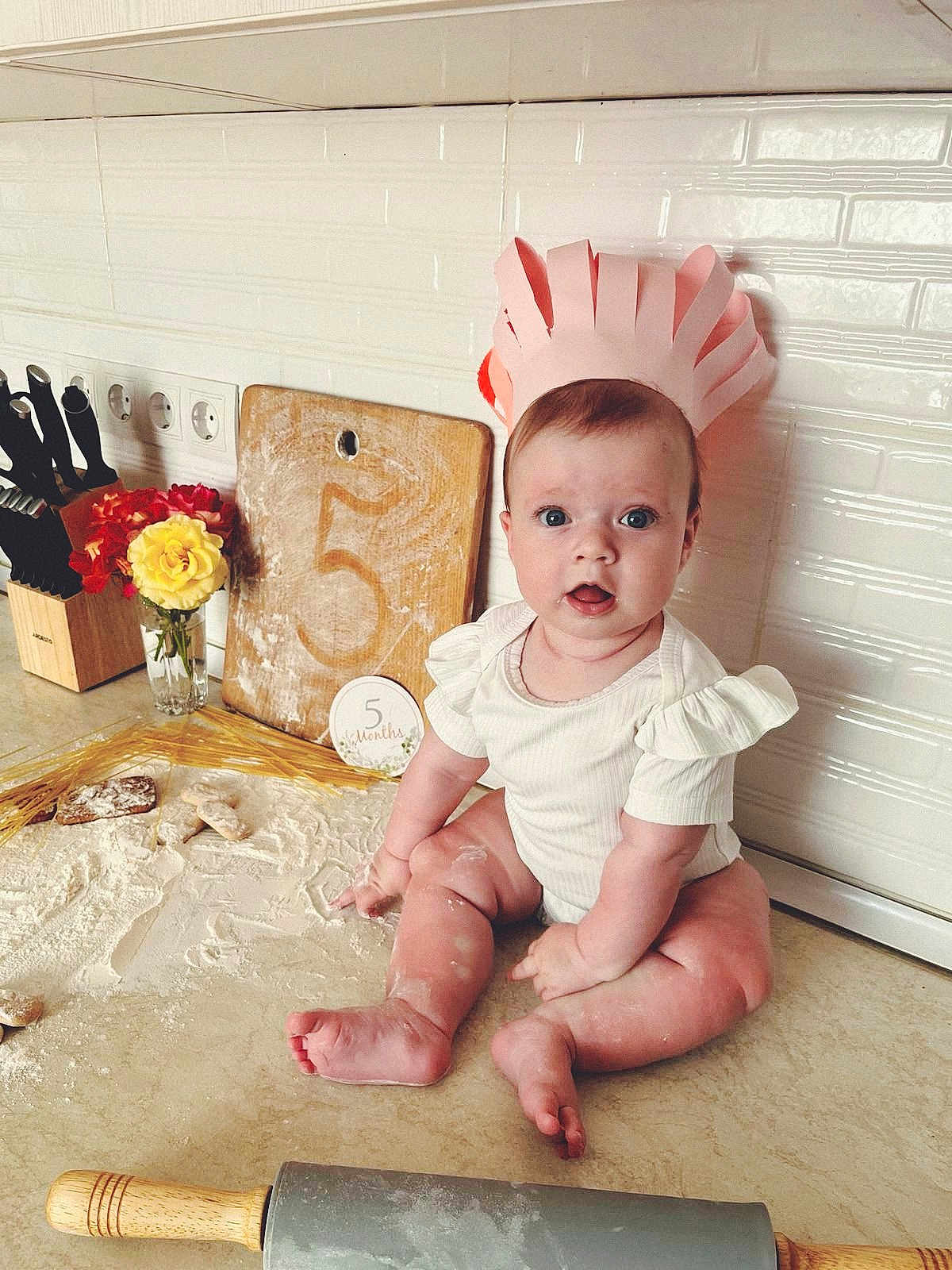 Solomia participe au concours pour gagner de l'argent avec cette photo : baby, infant, paper_crown, kitchen_counter, flour, rolling_pin, cutting_board, flowers, knife_block, white_wall_tiles, celebration, 5_months, surprised_expression, cute, sitting, cooking, home, child, portrait, kitchen