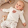 baby, infant, child, lying_down, white_clothing, animal_print, floral_pattern, wooden_toy, teething_ring, soft_surface, beige_background, cute, innocent, gazing_up, cozy, comfortable, indoor, portrait, young_child, peaceful