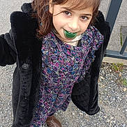 Amel participe au concours pour gagner de l'argent avec cette photo : child, girl, outdoor, coat, dress, smile, pacifier, brown_shoes, gravel, metal_railings, long_hair, playful, casual, patterned_clothing, standing, young, person, daylight, urban, happy