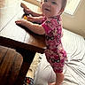 toddler, child, indoors, wooden_table, pink_onesie, snack, carpet, cushion, slipper, window_blinds, sunlight, smiling, person, standing, footwear, playful, casual, home, cute, happy