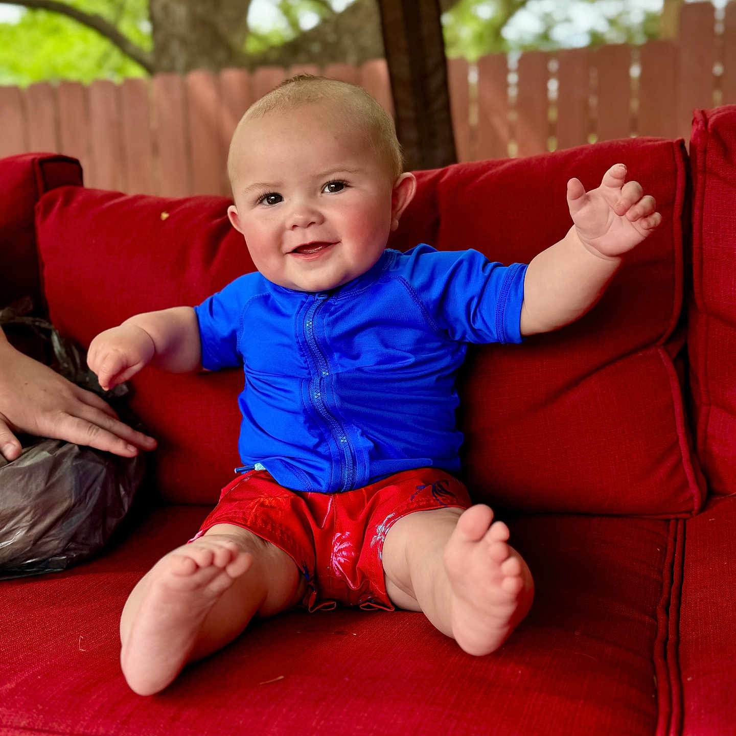Jaxon is registered to the contest to win money with this photo: baby, child, smiling, red_couch, blue_shirt, red_shorts, barefoot, outdoor, fence, tree, person, sitting, happy, cute, infant, portrait, hand, foot, skin, cushion