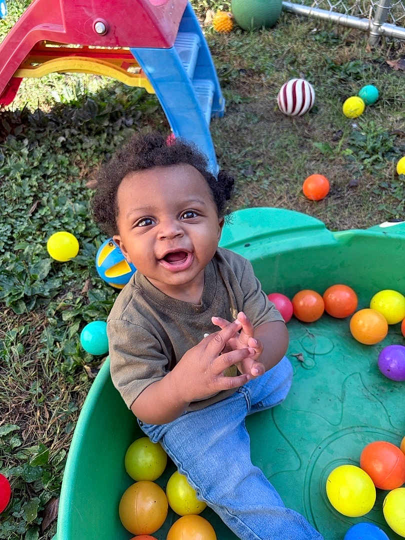 Messiah joined the competition — help win amazing prizes! toddler, child, smiling, outdoor, grass, plastic_container, colorful_balls, playground_slide, curly_hair, happy, casual_clothing, jeans, short_sleeve_shirt, play, fun, daylight, greenery, fence, toy, nature