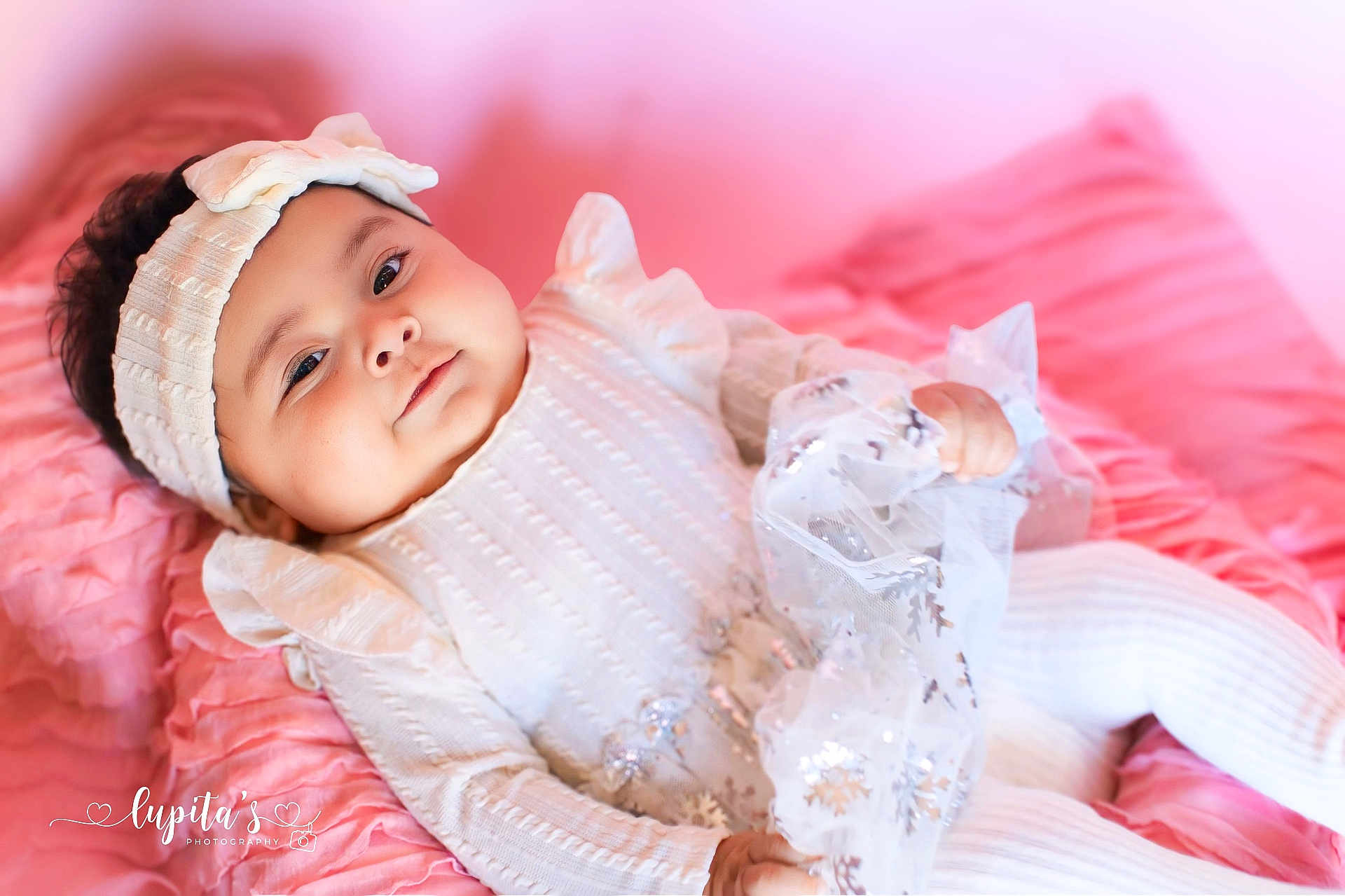 Alora joined the competition — help win amazing prizes! baby, infant, headband, bow, white_clothing, blanket, pink_background, fabric, snowflake_pattern, soft_lighting, portrait, cute, child, lying_down, cozy, indoors, smiling, chubby_cheeks, hand, photography