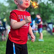 Brandon is registered to the contest to win money with this photo: child, sparkler, face_paint, red_shirt, black_shorts, glow_stick, outdoor, grass, summer, celebration, people, blurred_background, daylight, american_flag, old_navy, shorts, person, fun, festival, park