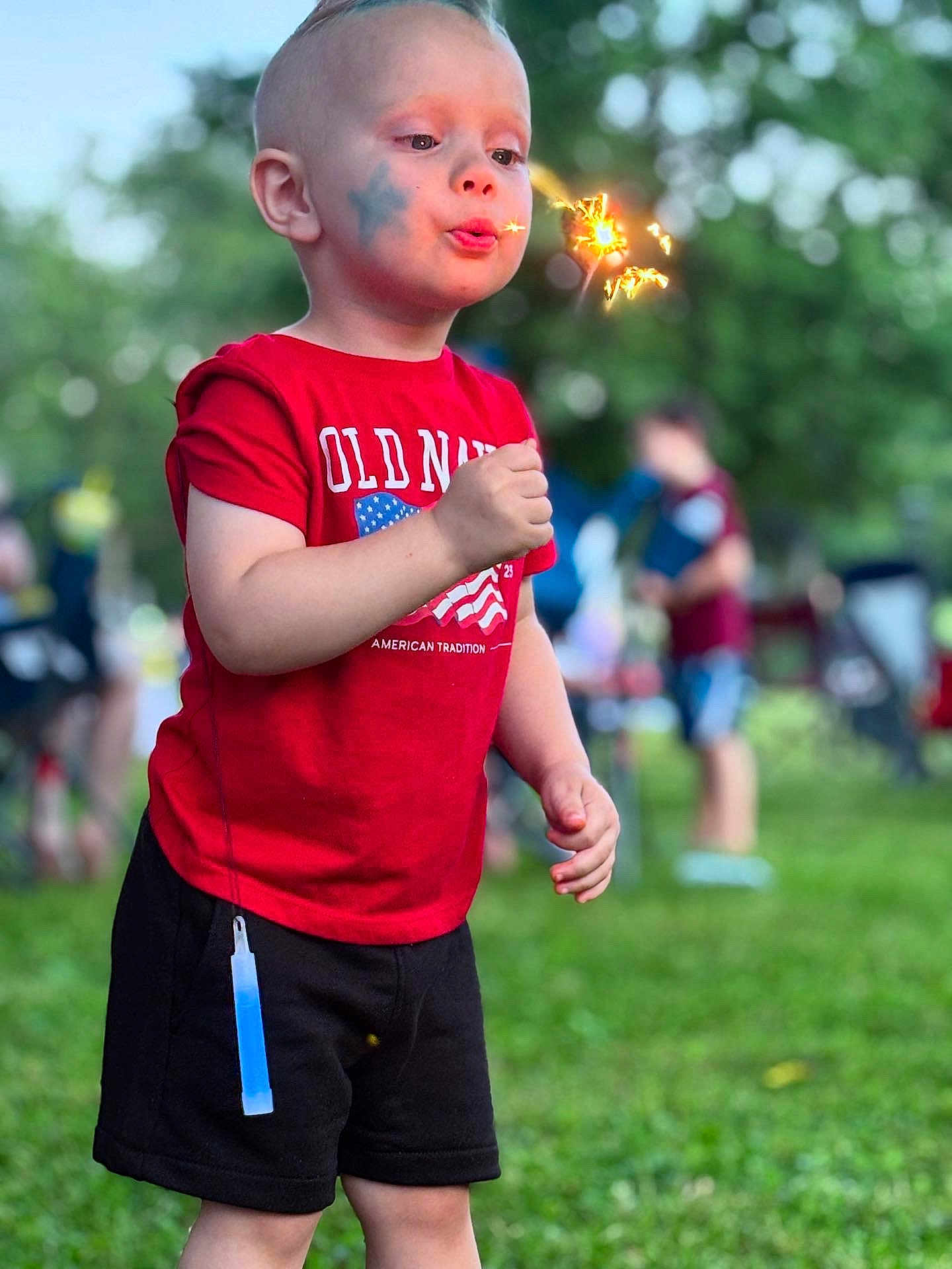 Brandon is registered to the contest to win money with this photo: child, sparkler, face_paint, red_shirt, black_shorts, glow_stick, outdoor, grass, summer, celebration, people, blurred_background, daylight, american_flag, old_navy, shorts, person, fun, festival, park