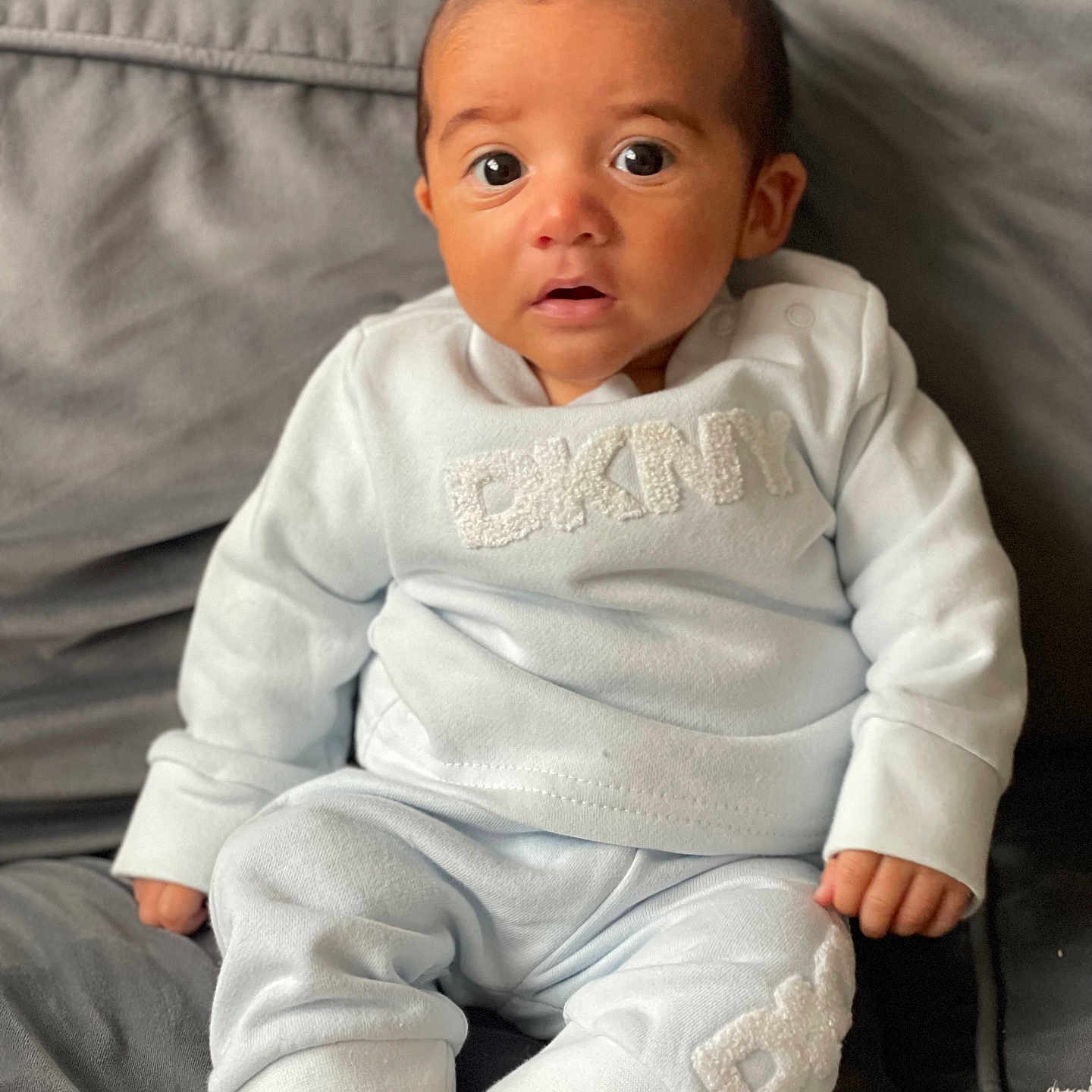 Vinnie-James is registered to the contest to win money with this photo: apparel, baby, blue_outfit, child, clothing, couch, cute, expression, eyes, face, feet, hands, home, indoors, infant, person, portrait, sitting, soft_fabric, young
