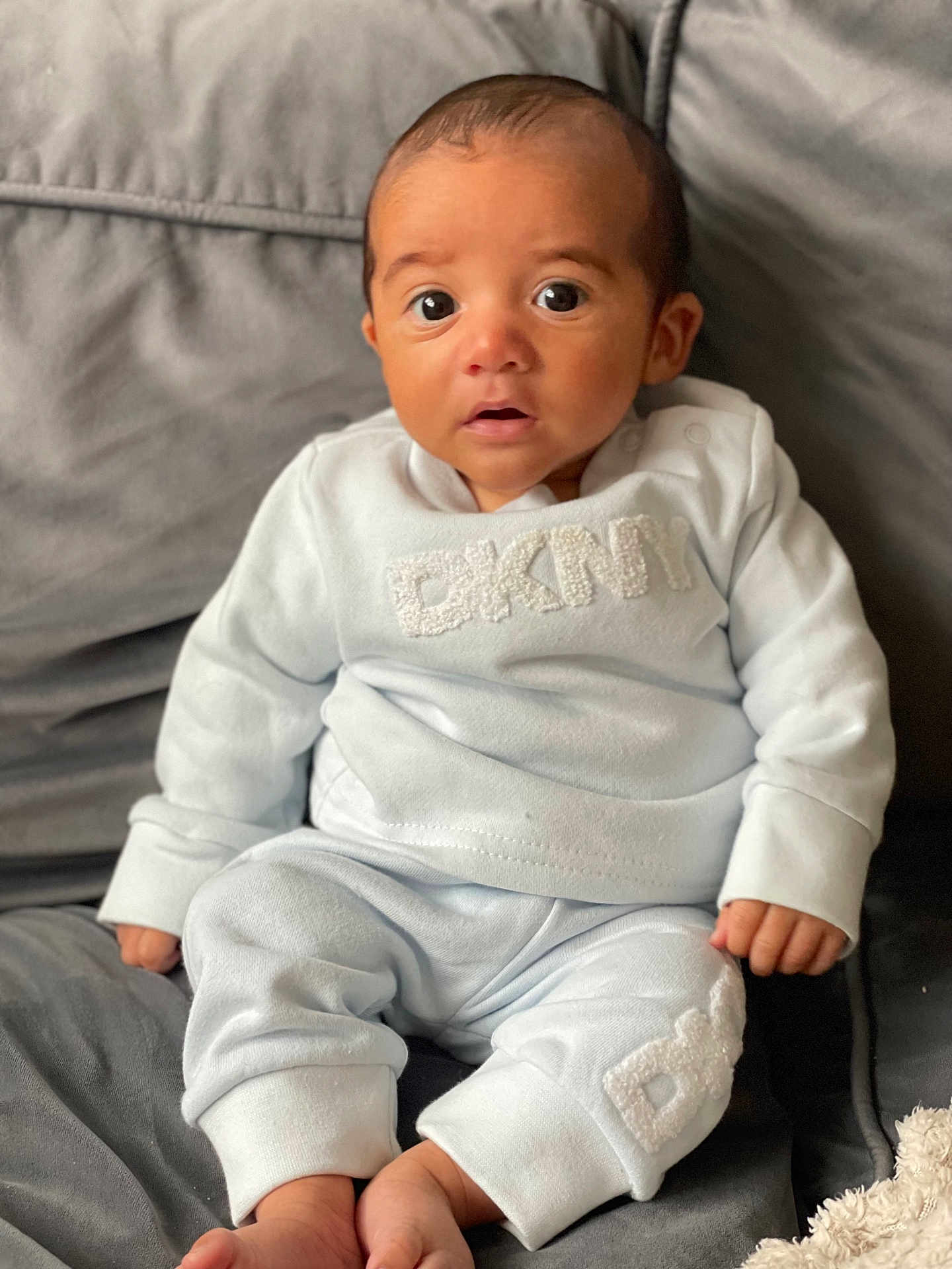 Vinnie-James is registered to the contest to win money with this photo: baby, infant, child, couch, clothing, blue_outfit, face, eyes, expression, sitting, hands, feet, home, indoors, soft_fabric, cute, portrait, young, person, apparel