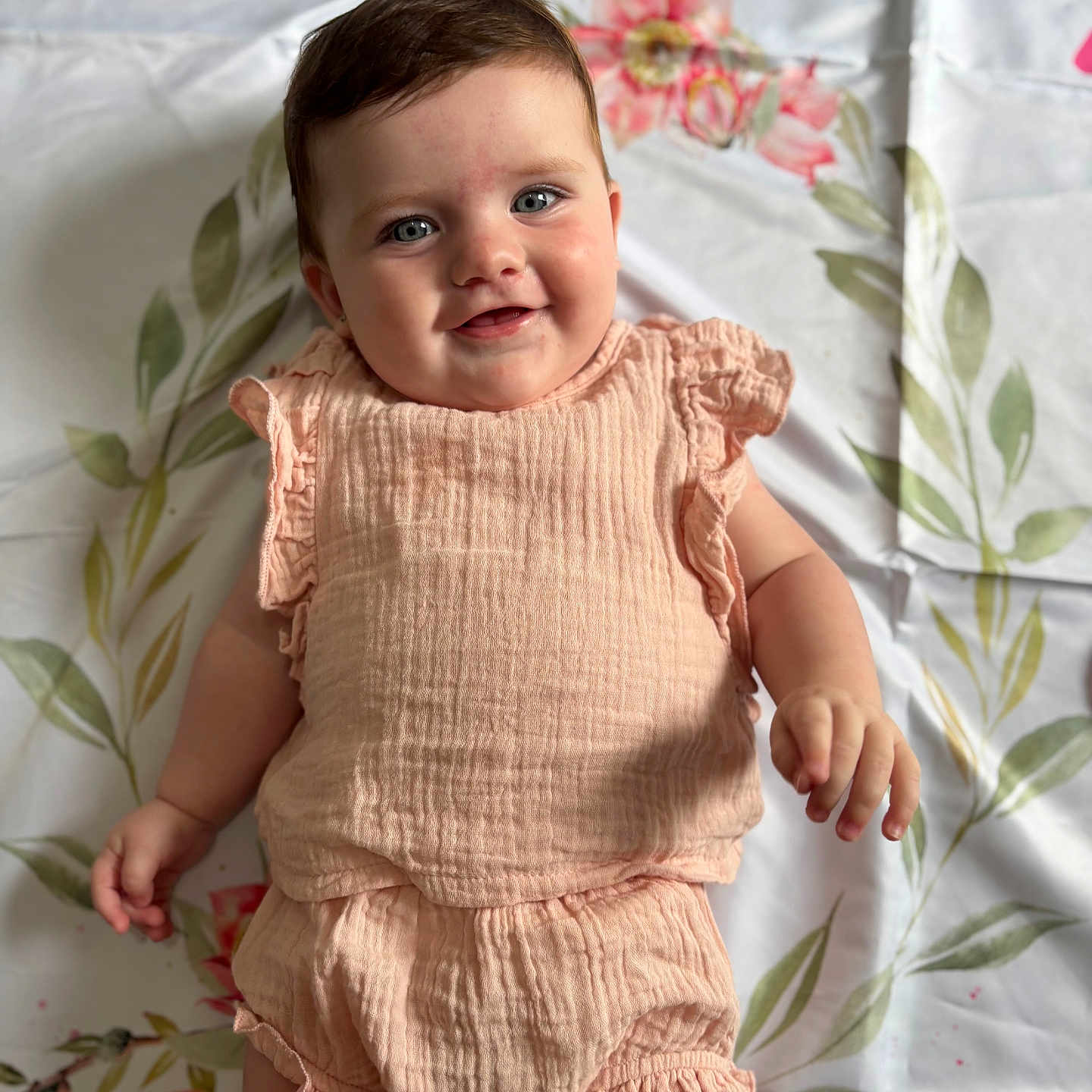 Arabella is registered to the contest to win money with this photo: baby, blue_eyes, child, chubby, cute, face, floral_blanket, hand, happy, indoor, infant, lying_down, peach_outfit, person, portrait, ruffles, skin, smiling, soft_lighting, young