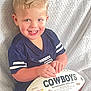 child, toddler, boy, smile, football, jersey, cowboys, ball, sports, portrait, blond_hair, blue_eyes, hands, seated, fabric_backdrop, indoor, happy, cute, closeup, toy