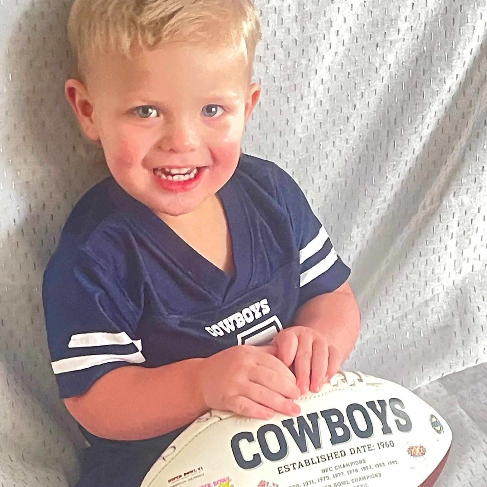 Witten is registered to the contest to win money with this photo: ball, blond_hair, blue_eyes, boy, child, closeup, cowboys, cute, fabric_backdrop, football, hands, happy, indoor, jersey, portrait, seated, smile, sports, toddler, toy