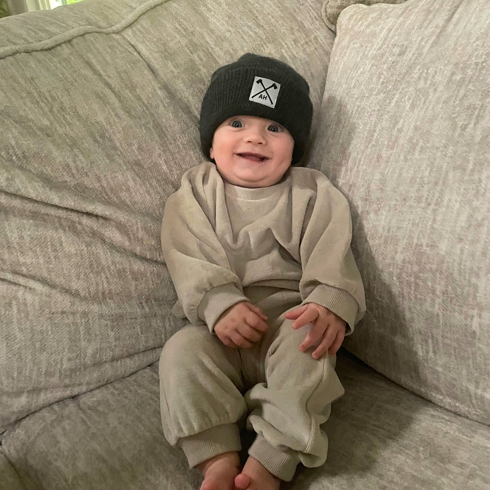 Emmett is registered to the contest to win money with this photo: baby, baby_toddler_clothing, baseball_cap, cap, comfort, eye, face, foot, grey, happy, head, headwear, joy, linens, outerwear, pattern, person, sitting, sleeve, smile