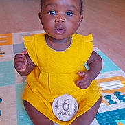 Micka participe au concours pour gagner de l'argent avec cette photo : baby, child, yellow_dress, bows, play_mat, sitting, indoor, wooden_plaque, age_marker, curious, cute, infant, portrait, floor, skin, hands, feet, bright_colors, expression, adorable