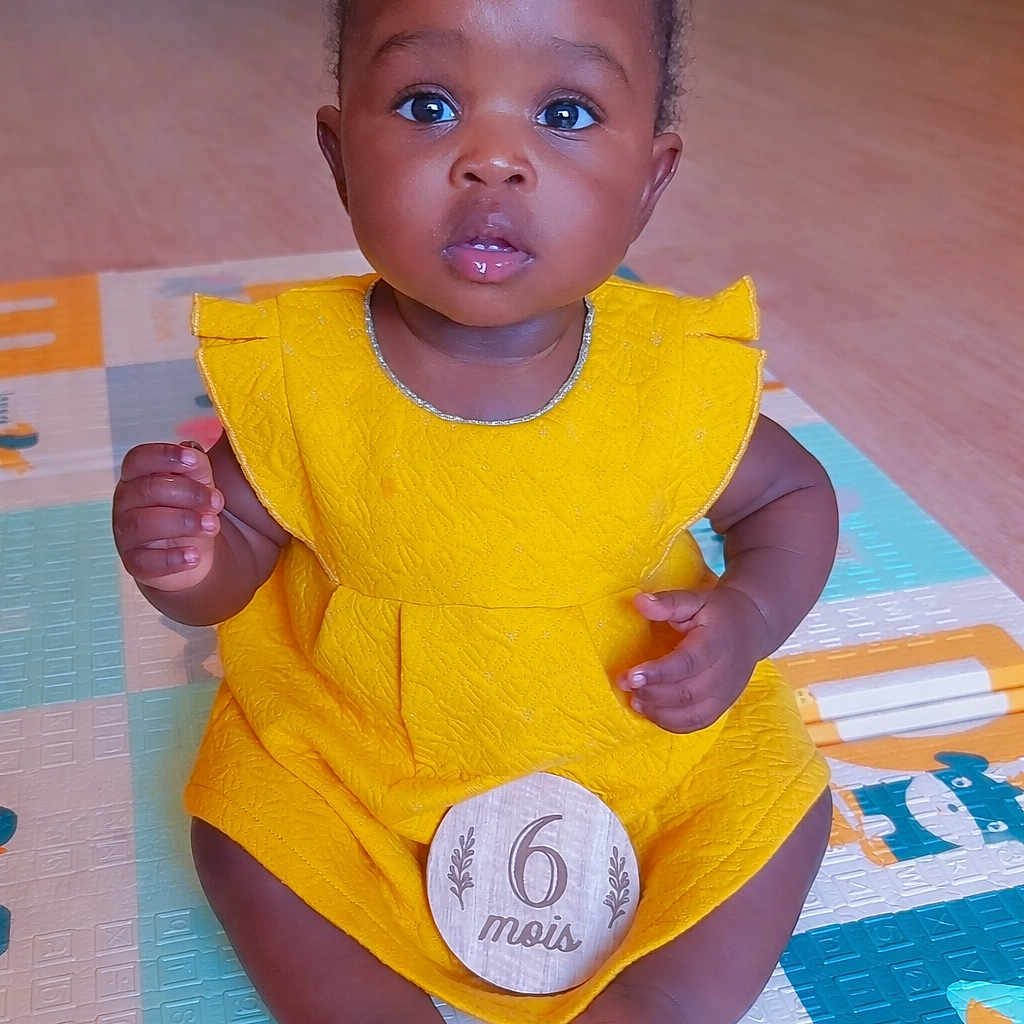 Micka participe au concours pour gagner de l'argent avec cette photo : baby, child, yellow_dress, bows, play_mat, sitting, indoor, wooden_plaque, age_marker, curious, cute, infant, portrait, floor, skin, hands, feet, bright_colors, expression, adorable
