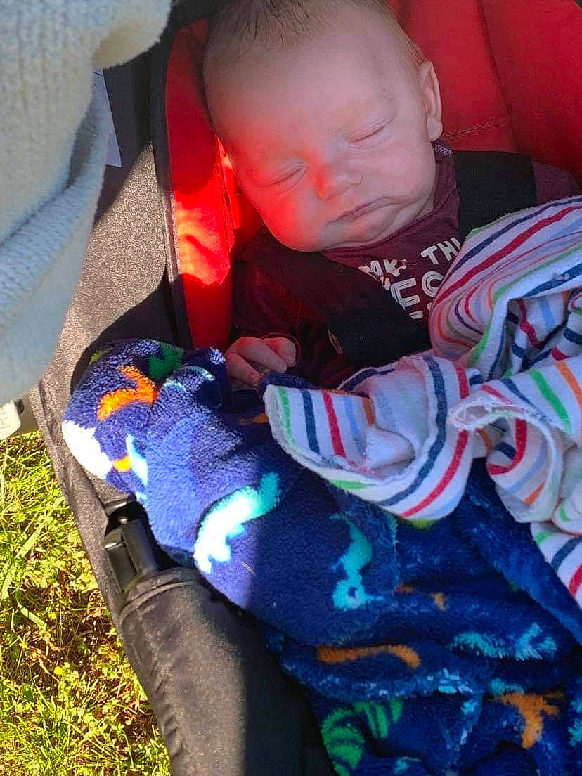 Kyle is registered to the contest to win money with this photo: baby, baby_products, baby_toddler_clothing, car_seat, carmine, child, comfort, electric_blue, grass, linens, pattern, person, personal_protective_equipment, sitting, sleeve, toddler, vertebrate