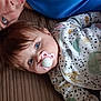 adult_face, animal_print, baby, blue_eyes, brown_background, child, close_up, cute, family, head, human, indoors, infant, lying_down, onesie, pacifier, person, portrait, sleepwear, soft_lighting