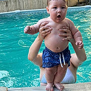 Brody is registered to the contest to win money with this photo: adult_hands, baby, child, expression, family, fun, holding, outdoor, person, playful, pool_edge, recreation, skin, splash, summer, sunlight, swim_trunks, swimming_pool, water, wet_hair
