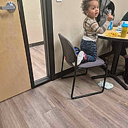 Kingston is registered to the contest to win money with this photo: toddler, child, indoor, chair, table, snack, sippy_cup, jeans, curly_hair, sneakers, backpack, wood_floor, door, light_switch, person, casual_clothing, expression, kneeled, room, furniture