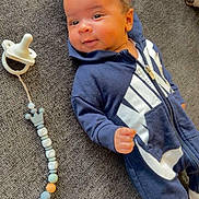 Truu' is registered to the contest to win money with this photo: baby, infant, newborn, onesie, hoodie, pacifier, pacifier_clip, zipper, lying_down, couch, gray_texture, carpet, face, eyes, smile, hand, hair, cozy, portrait, relaxed