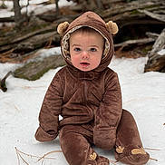 Beau is registered to the contest to win money with this photo: baby, child, snow, forest, brown_onesie, hood, cute, drool, sitting, winter_clothing, outdoor, nature, logs, pine_needles, eyes, face, warm_clothes, fur_texture, footwear, hood_ears