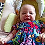 baby, child, smiling, floral_dress, colorful_clothing, infant, cushion, baby_seat, indoor, happy, person, face, skin, head, arm, hand, fabric, pattern, comfort, cute