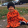 child, toddler, smiling, orange_outfit, pumpkin_pattern, black_beanie, sitting, sidewalk, grass, autumn, leaves, parked_cars, residential_buildings, outdoor, daylight, casual_clothing, happy, cute, person, footwear