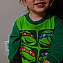 child, pajamas, green_clothing, teenage_mutant_ninja_turtles, person, face, hand, indoor, sitting, wall, power_outlet, expression, young_child, looking_up, furniture, adult_hand, brown_hair, casual, home, clothing