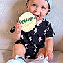 baby, child, infant, smiling, cap, backward_hat, black_clothing, lightning_bolt_pattern, sitting, holding_sign, seven_months, white_shoes, soft_surface, cute, blue_eyes, tongue_out, portrait, indoor, young_child, happy