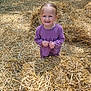 child, girl, purple_clothing, straw, hay, outdoor, field, sunlight, nature, standing, person, smile, expression, grass, daytime, casual, cute, young, portrait, playful