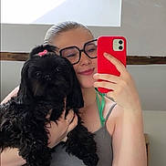 Kiki a rejoint le concours — aidez-le/la à gagner de superbes lots ! selfie, woman, dog, black_dog, glasses, pink_bow, smartphone, red_phone_case, indoor, casual_clothing, smile, long_hair, holding_pet, pet_owner, portrait, mirror, window, natural_light, happy, cute_pet