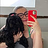 selfie, woman, dog, black_dog, glasses, pink_bow, smartphone, red_phone_case, indoor, casual_clothing, smile, long_hair, holding_pet, pet_owner, portrait, mirror, window, natural_light, happy, cute_pet