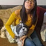Zuma is registered to the contest to win money with this photo: woman, glasses, dog, couch, indoor, yellow_top, jeans, long_hair, pet, small_dog, bow, casual, seated, brown_couch, cushion, home, relaxed, portrait, friendly, furniture