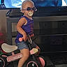 Oaklynn joined the competition — help win amazing prizes! toddler, child, toy_bike, indoor, sunglasses, pacifier, purple_shirt, shorts, television, subtitles, floor, black_and_white_tiles, plastic_toy, furniture, entertainment_center, person, cute, playtime, casual, helmet_absent