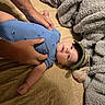 baby, child, infant, portrait, lying_down, bed, blanket, fuzzy_blanket, blue_outfit, green_hat, cap, adult_hands, family, home, indoor, smiling, wide_eyed, cute, cozy, photography