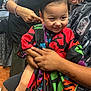 child, haircut, barber, barbershop, cape, scissors, person, smile, happy, hair, adult, hand, tattoo, colorful, indoor, clothing, hairstyle, arm, sitting, people