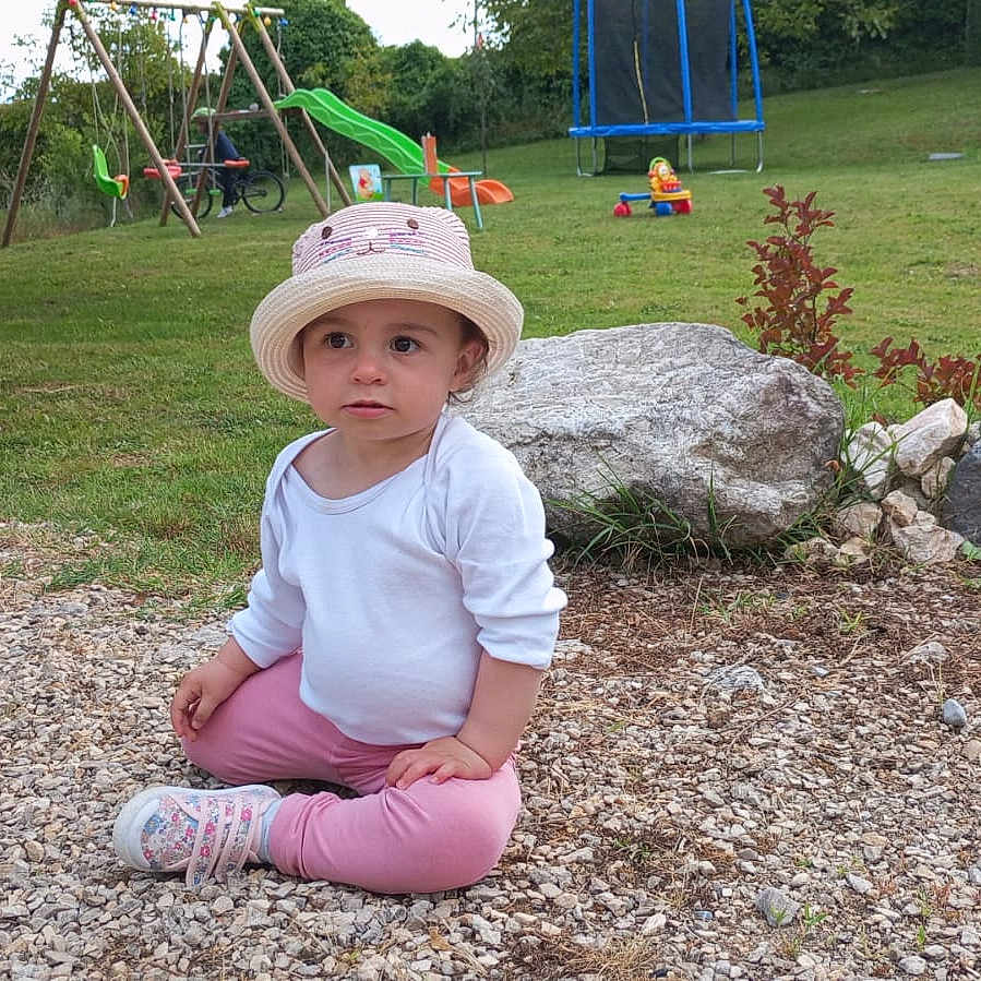 Jade a rejoint le concours — aidez-le/la à gagner de superbes lots ! casual_clothing, child, garden, grass, gravel, green_slide, hat, leisure, nature, outdoor, pink_pants, play_equipment, playground, rocks, shoes, sitting, toddler, trampoline, white_shirt, young_child