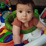 William is registered to the contest to win money with this photo: baby, child, infant, portrait, face, eyes, wide_eyed, sitting, activity_center, jumper, baby_toys, colorful, indoor, home, brown_hair, short_sleeves, green, orange, white, curious