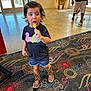 child, toddler, lollipop, indoor, carpet, cowboy_boots, denim_shorts, black_tshirt, person, curious_expression, colorful, pattern, footwear, short_hair, playful, walking, background, door, plant, casual_clothing