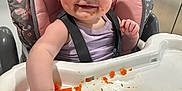 Maci is registered to the contest to win money with this photo: baby, high_chair, food, messy, kitchen, child, smiling, face, hand, cushion, purple_clothing, feeding, orange_food, tray, indoor, person, happy, hair, seatbelt, table
